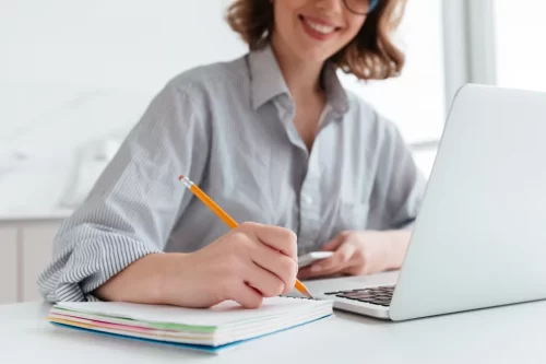 young-smiling-woman-in-striped-shirt-taking-notes-while-sitting-at-table-in-light-apartment